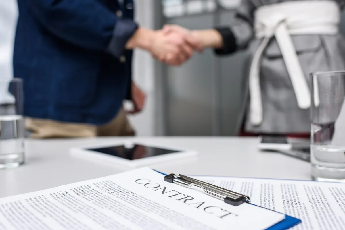 Business partners shaking hands in an office setting with a signed contract on a clipboard in the foreground, symbolizing agreement and collaboration.