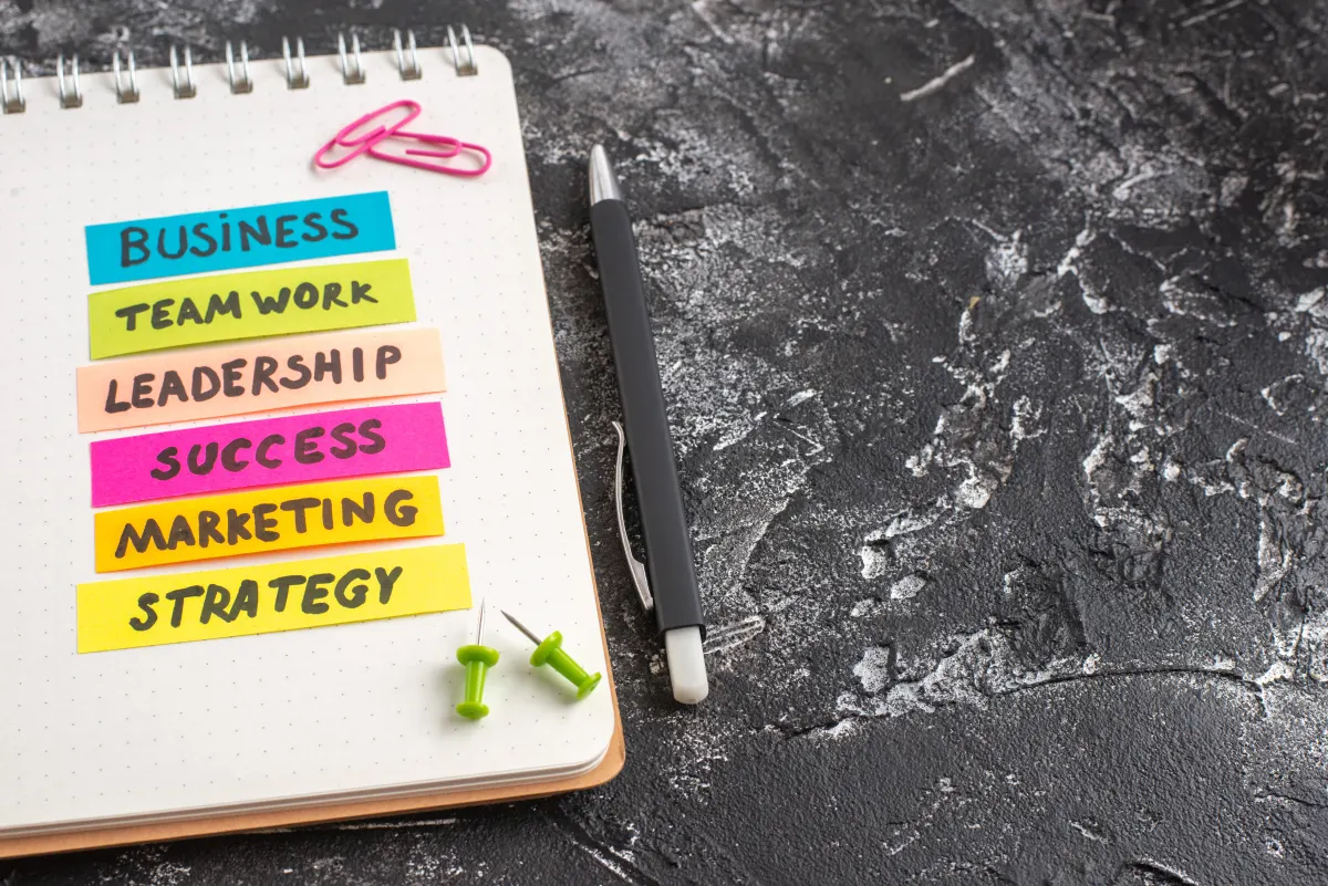 Colorful sticky notes on a notebook displaying different business terms, with a pen and push pins on a textured black surface.