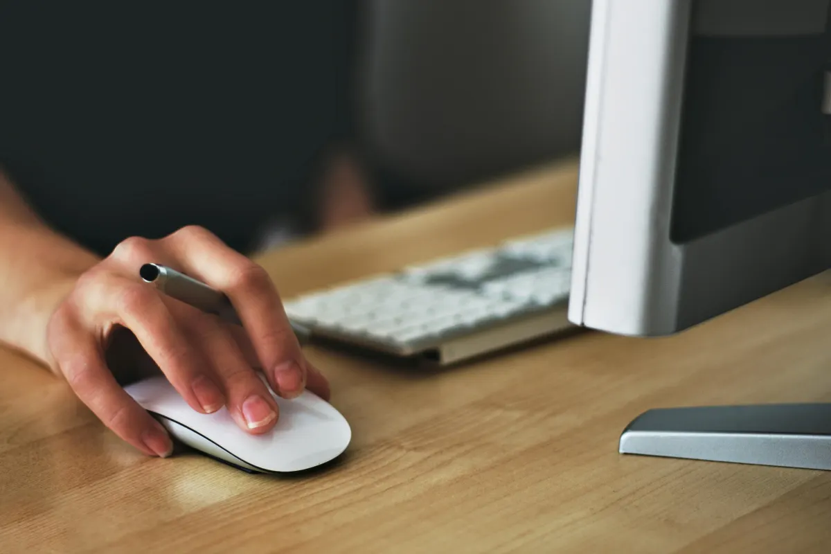 Hand holding a white computer mouse on a wooden desk with a keyboard and monitor visible