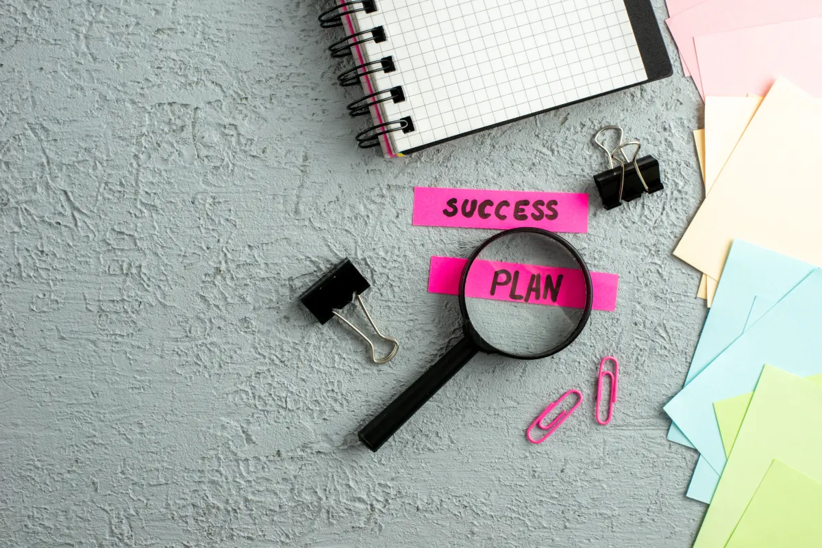 op view of a notebook, colorful envelopes, paper clips, binder clips, and a magnifying glass highlighting the words "sucess" and "plan" on pink sticky notes.