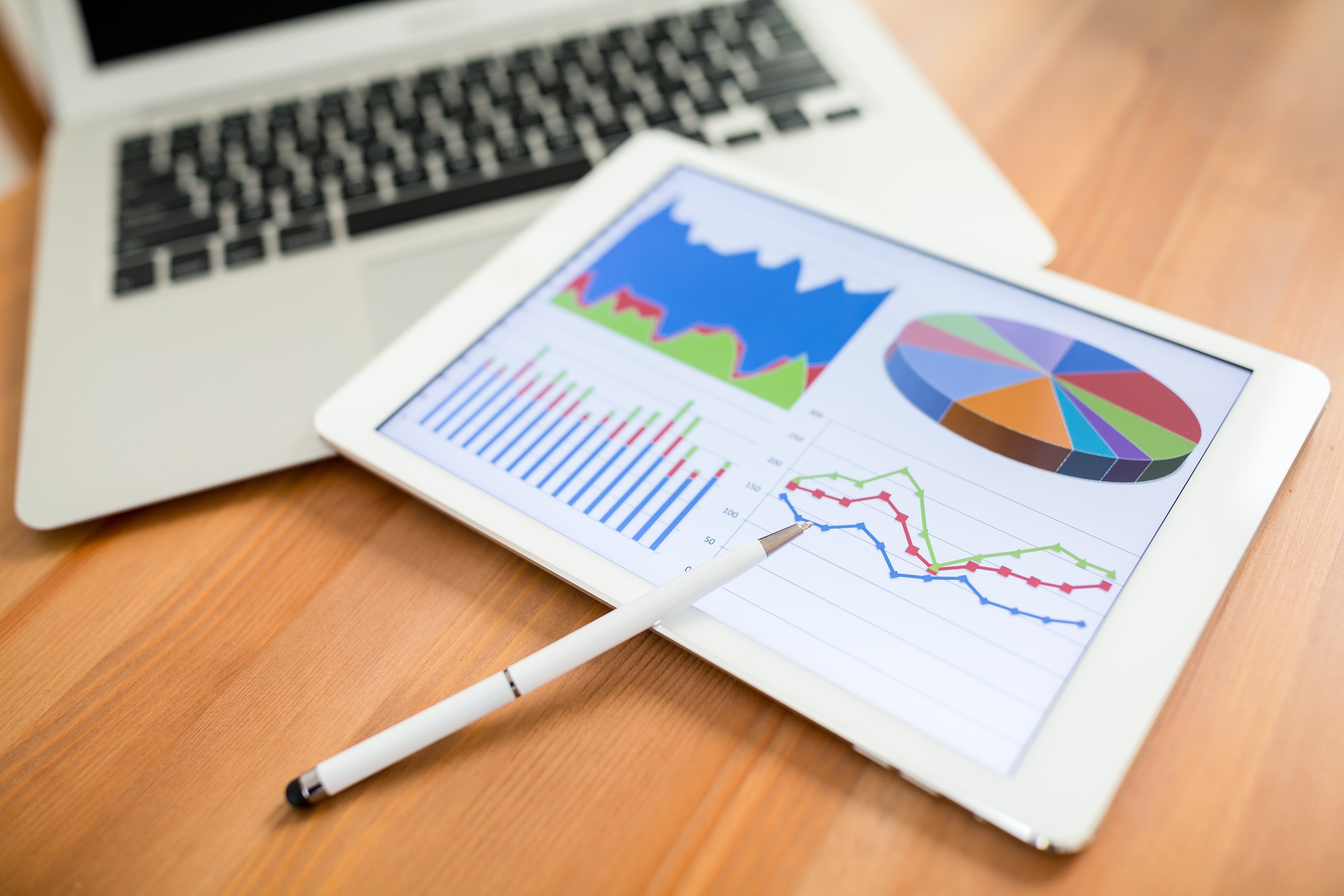 Tablet displaying business graphs and charts, including a bar graph, pie chart, and line graphs, with a stylus pen on a wooden desk near a laptop.