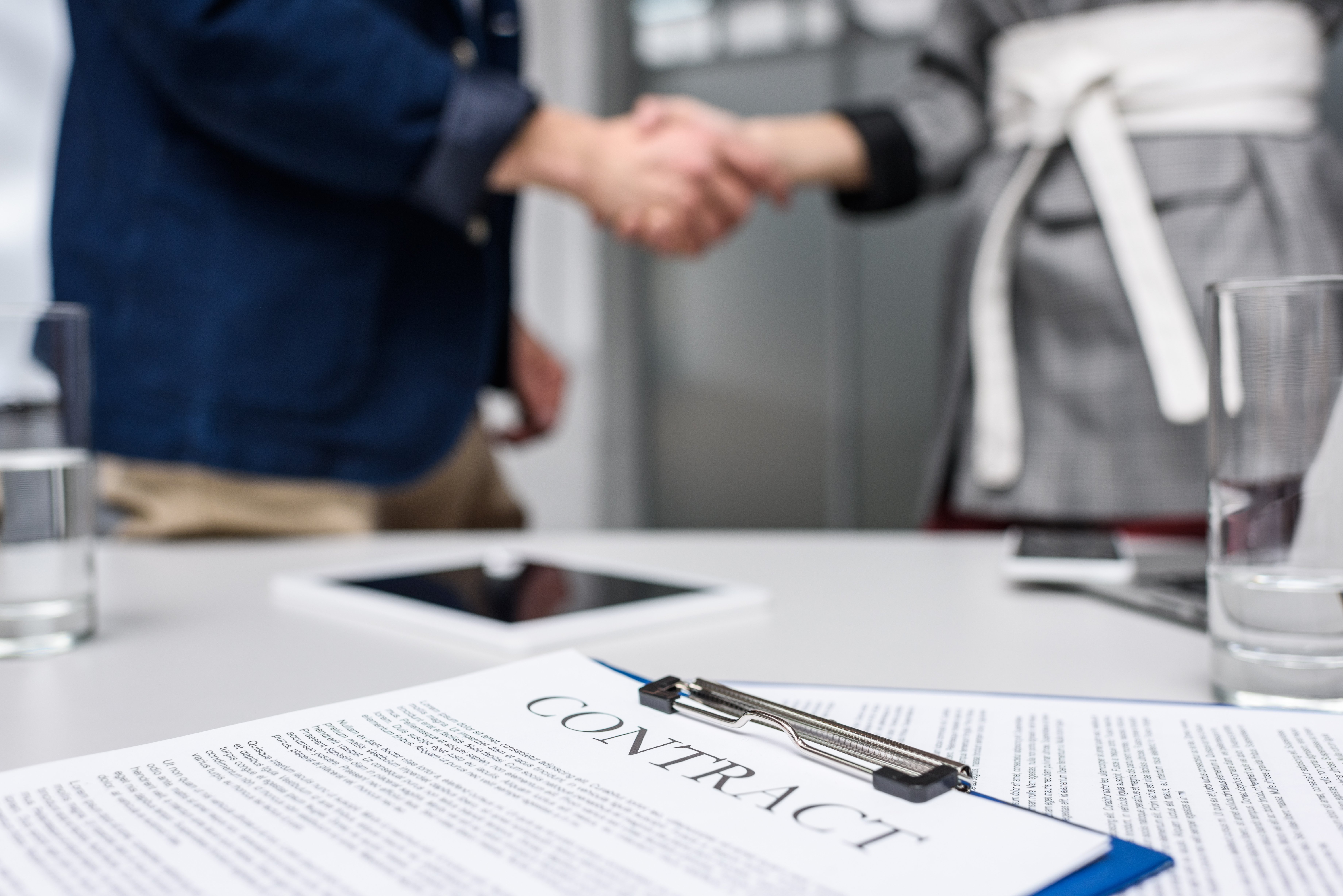 Business partners shaking hands in an office setting with a signed contract on a clipboard in the foreground, symbolizing agreement and collaboration.