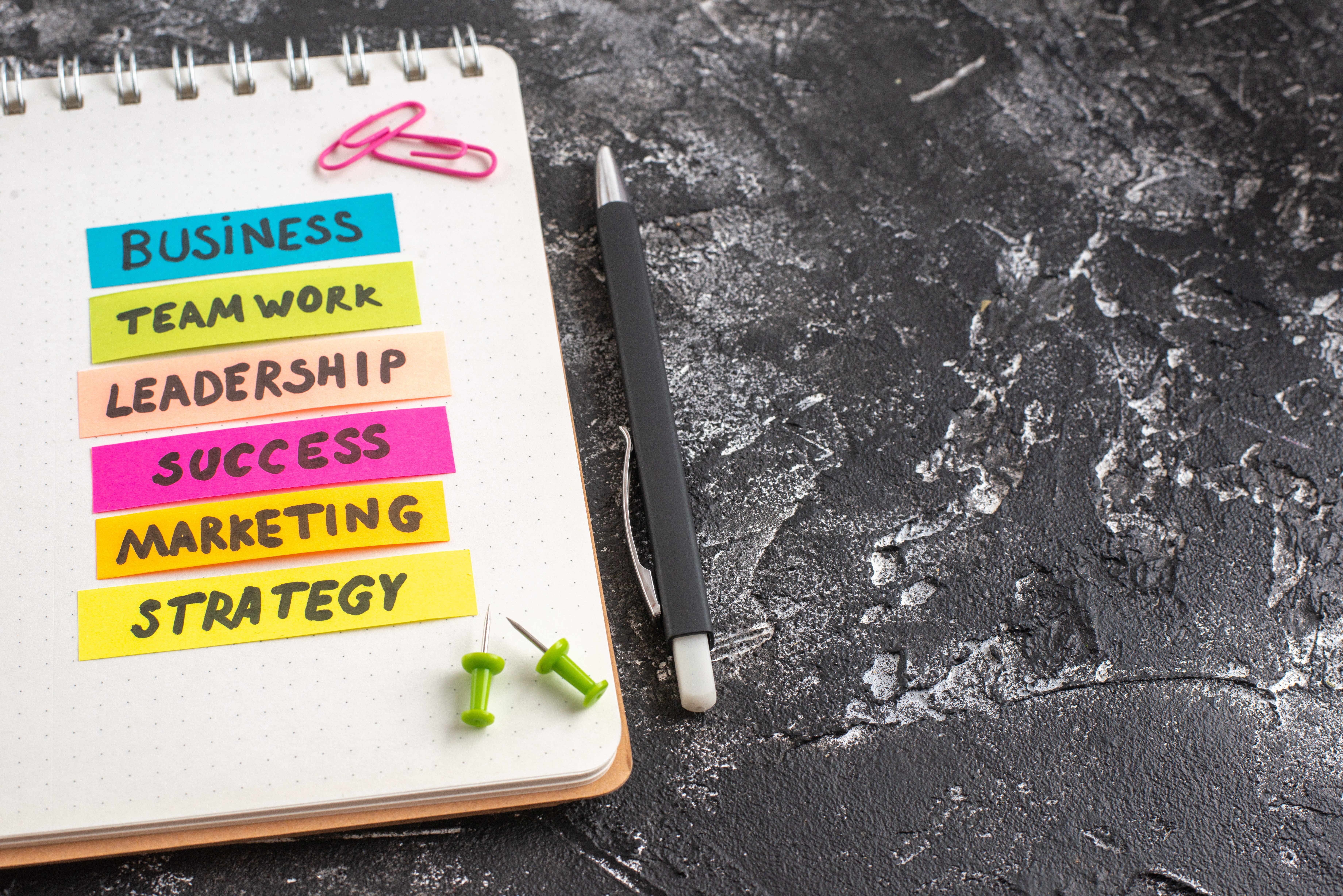 Colorful sticky notes on a notebook displaying different business terms, with a pen and push pins on a textured black surface.