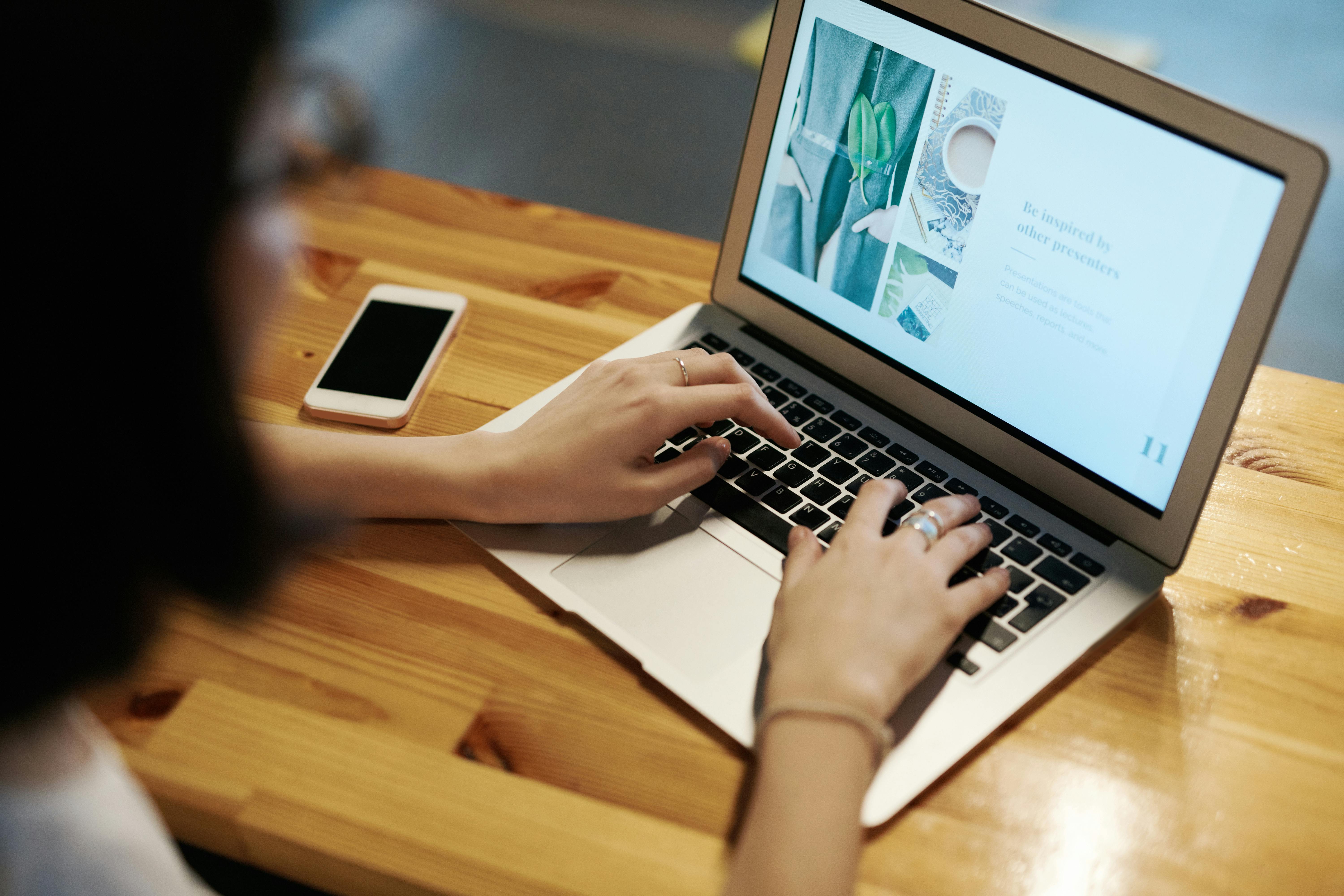 Person typing on a laptop with a design presentation on screen and a smartphone nearby.