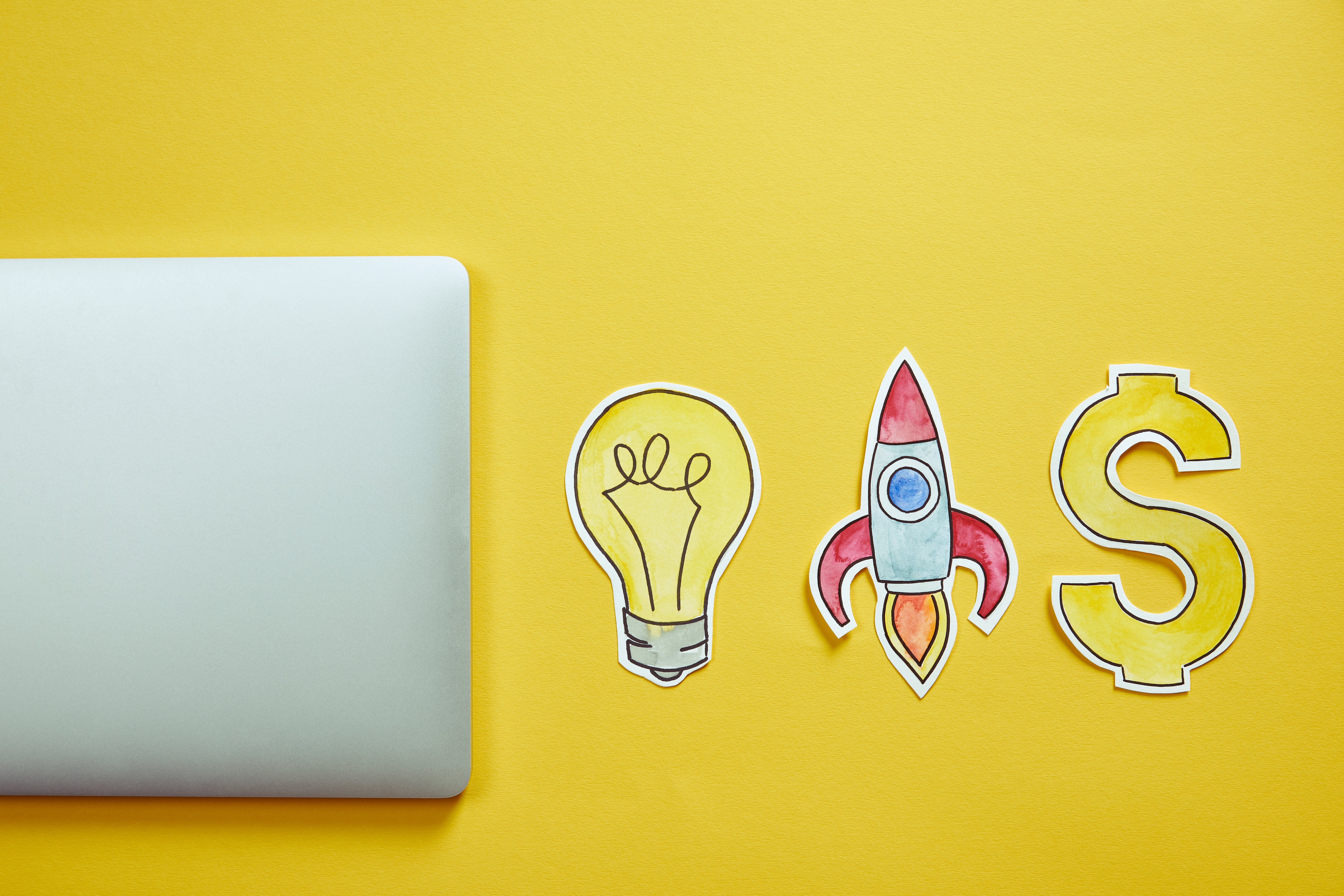 Top view of a laptop and hand-drawn paper cutouts of a lightbulb, a rocket, and a dollar sign on a yellow background, symbolizing innovation, growth, and financial success.